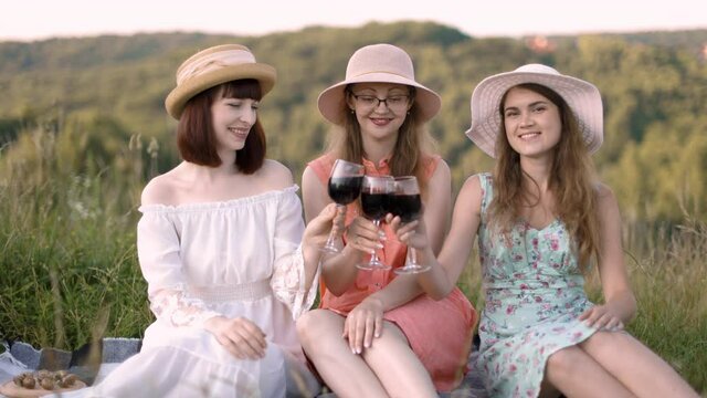 Cheerful female friends have a great time together on a picnic in a picturesque meadow with green hills. Girls in hats and dresses sitting on the blanket and toasting wine glasses