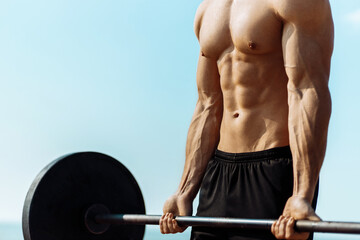 Muscular man training on the beach by the sea, doing exercises with a barbell for biceps, strong...