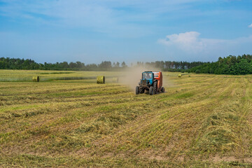 Obraz premium On the rye field, the tractor is harvesting in fresh grass hay.