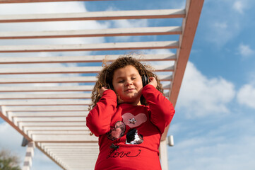 Little girl with curly hair wearing headphones, listening to music with closed eyes in public park....