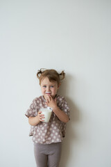  a little girl with tails against a white wall is standing in her hands holding a glass of kefir dissatisfied                              