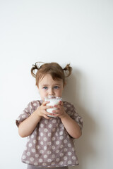 a little girl with tails against a white wall is standing in her hands holding a glass of kefir...