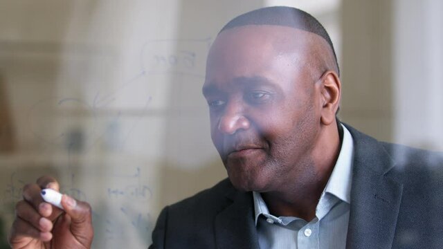View through glass mature middle aged african business man male wrinkled focused face professor specialist professional scientist writes on transparent board equation counts profit finance development