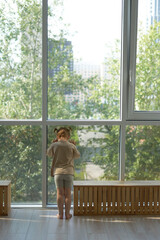 a little girl in light clothes is standing at home looking out of a stained glass window at the street with sadness