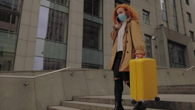 Young red-haired woman going with yellow suitcase and talks on the phone in protective face mask. Safe travel. High quality 4k footage
