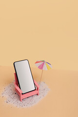 A phone with a white blank screen in a pink beach foldable deck chair standing on sand surface next to a cocktail sun umbrella against beige background. Creative summer concept with a copy space.