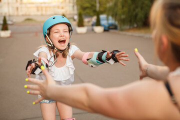 Excited young girl wearing protective helmet and gear, learning rollerblading, skating to hug her mother