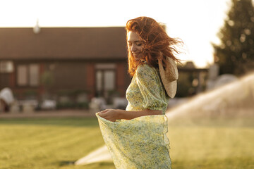 Red haired fashionable young lady straw modern hat in trendy green and yellow dress smiling with closed eyes and posing outdoor..