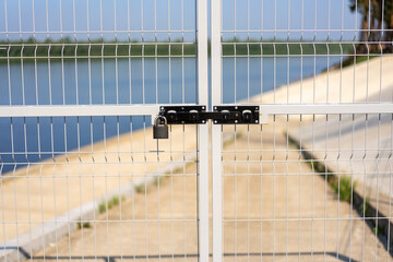 The gate with a metal vertical lattice fence is closed with a padlock on the background of the river