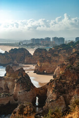 As the sun goes down natural rock archs and cliffs form a scenic view of the beach, a city is visible in the background