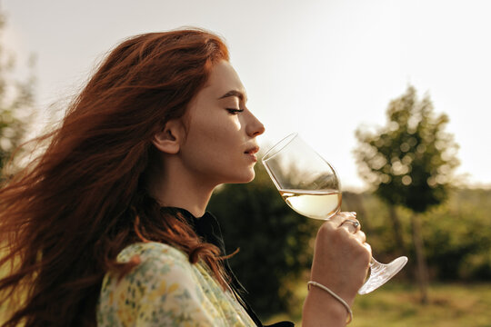 Profile photo of red haired young woman in stylish green clothes and bracelet holding glass with champagne and drinking outdoor..