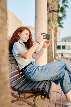 Young redhead woman takes a picture with her mobile sitting in a bench