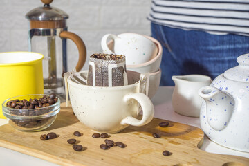 Girl hands in the frame, close-up, make instant freshly brewed cup of coffee from bag. Trendy variety of drip coffee, in a bright kitchen on a table with cups, morning sun light