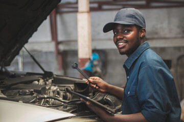 Fototapeta na wymiar African maintenance male checking car with use wrench and tablet , service via insurance system at Auomobile repair and check up center