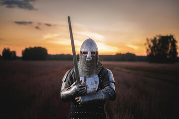 Knight guardian in plate armor with a sword in hands stands in the field on a sunset background. The kingdom defender.
