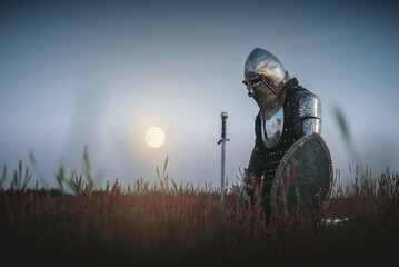 The tired knight in the plate armor kneels among the battlefield with a sword in the moon light. Lost battle. Defeat concept. - obrazy, fototapety, plakaty