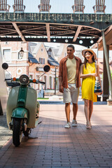 stylish multiethnic couple looking at scooter while walking on urban street