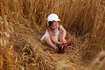 Naklejka na ściany i meble a beautiful cute little blonde girl drinks milk from a clay mug there is a clay jug next to her in a wheat field at sunset