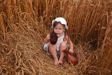 a beautiful cute little blonde girl drinks milk from a clay mug there is a clay jug next to her in a wheat field at sunset