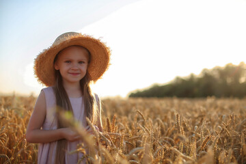 a beautiful blonde girl with long hair in a pink linen dress and a straw hat stands in a wheat field in the setting sun