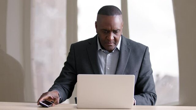Concentrated mature middle age african american business man manager boss leader sitting at table in office working at computer online with laptop answering call talking on mobile phone smartphone