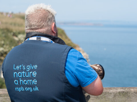 Bempton-Yorkshire-UK-July 17,2021 : A rear view of an RSPB warden looking out to sea at Bempton Nature Reserve.He wears a gilet with 'lets give nature a home ' and website printed on rear.Blue sky 