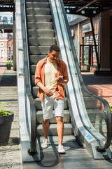 cheerful african american man in shorts using mobile phone near escalator