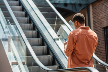 young african american man in shirt messaging on mobile phone on escalator - Powered by Adobe