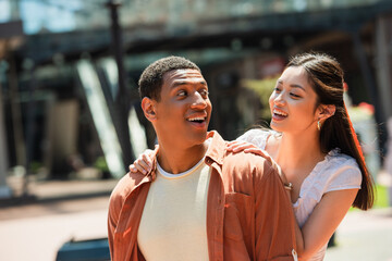 Obraz premium excited multiethnic couple smiling at each other while walking in city
