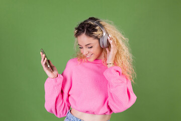 Young blonde woman with long curly hair in pink sweater on green background happy positive enjoying favorite  music in wireless  headphones