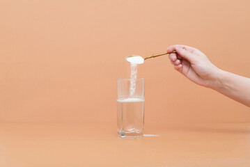 Hand pours collagen protein powder in a glass of water on a beige background. A natural supplement for skin beauty and bone health.
