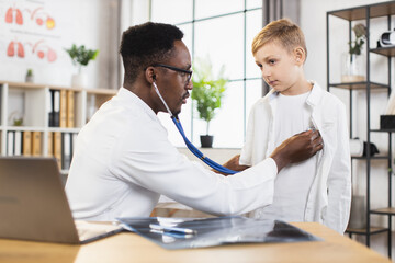 Pediatry, regular check up. Handsome skillful afro-american doctor checking boy's heartbeat and lungs breath during his visit to modern medical center. Close up shot.