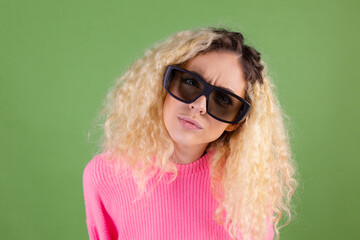 Young blonde woman with long curly hair in pink sweater on green background in 3d  cinema glasses serious focused look  to camera