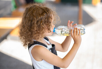 Little girl with curly hair drinks water from a bottle on the street on a summer day
