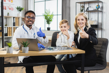 Healthcare concept. Handsome smiling professional afro-american doctor sitting at the table together with pretty joyful woman and her teen happy son gesturing thumbs up into camera in modern clinic