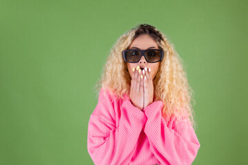 Young blonde woman with long curly hair in pink sweater on green background in 3d cinema shocked