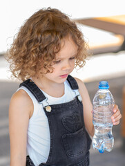 Little girl with curly hair drinks water from a bottle on the street on a summer day