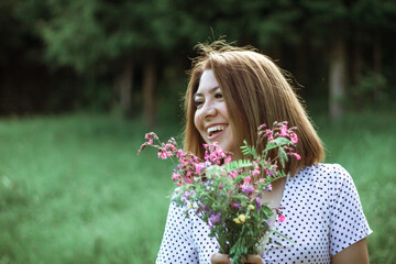 Fototapeta premium A girl with a short haircut and flowers in her hands looks away and laughs. A close portrait of a young beautiful woman in a meadow with lilac wildflowers in her hands. Selective focus. Copy space
