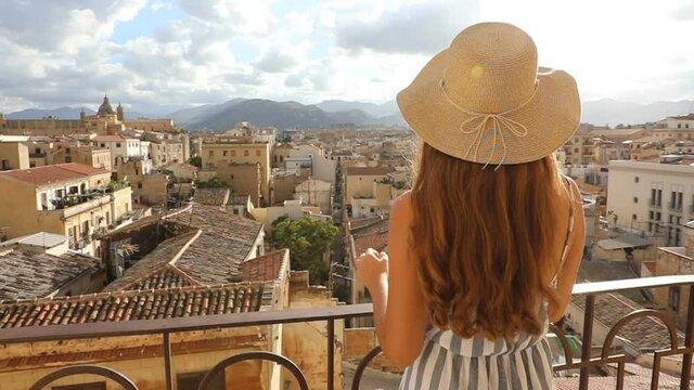 Attractive woman with hat on balcony looks out at cityscape of Palermo, Sicily, Italy