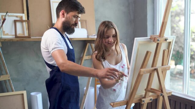 a couple of young artists working in an art studio creating a painting on canvas. The bearded artist teaches his girlfriend to paint canvas with paints, to use abstract works in the workshop.