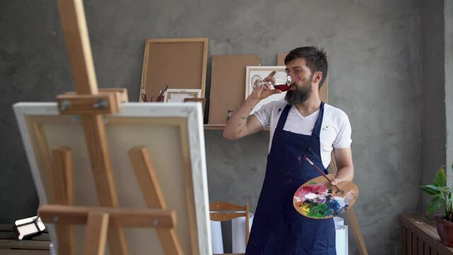 bearded man - an artist paints in an art workshop with a glass of wine and a palette in his hands. Painter is a thoughtful creative crisis. Lack of ideas. Creativity concept.