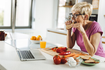 Senior woman eating a bread while reading online recipes in the kitchen