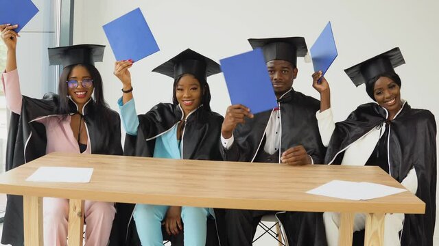 Four graduates of an African American university or college sit at a desk and show their blue diplomas