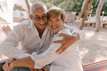 Positive man with gray mustache in eyeglasses and white shirt laughing and hugging his wife with...