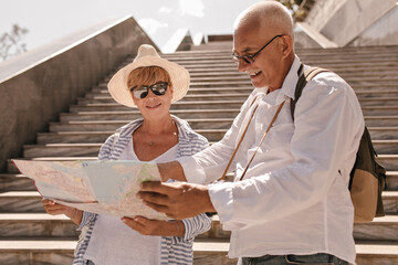 Grey haired man in glasses and light shirt with backpack looking at map with modern woman in hat...