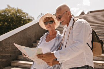 Happy man with grey hair in white shirt with backpack holding map and posing with blonde woman in blue striped clothes outdoor..