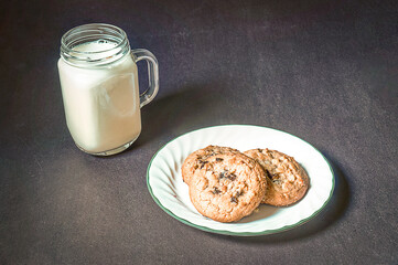 Cookies sit on a plate next to a glass of milk on a black background