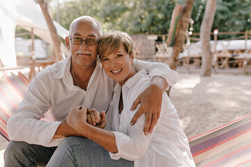 Wonderful woman with short blonde hairstyle in modern blouse smiling, sitting on hammock and hugging with her husband in eyeglasses on beach..