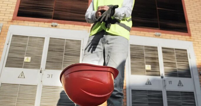 A power engineer puts on protective gloves on the background of an electrical substation, in the foreground an orange protective helmet. The use of personal protective equipment in production.
