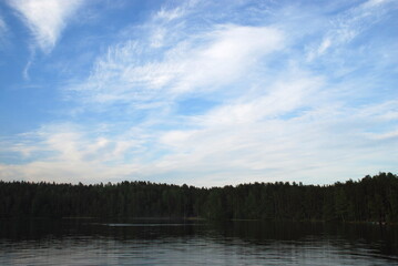 Light blue sky with white cirrus clouds. Almost calm surface of the lake with light ripple forest on the opposite shore under a light blue sky with clouds.